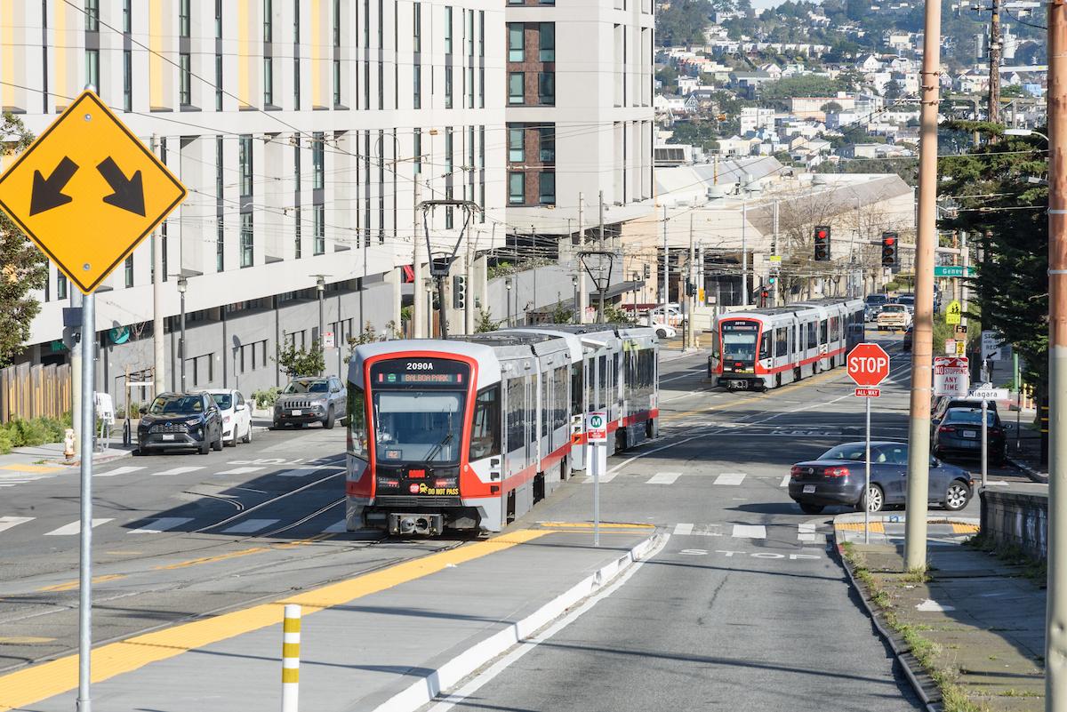 Light rail trains on a street with parked cars.