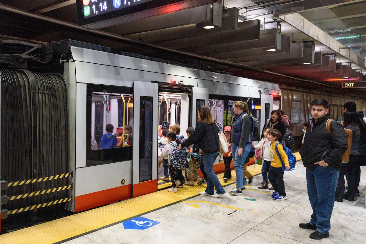 Adults and several students board a Muni Metro train from a station platform.
