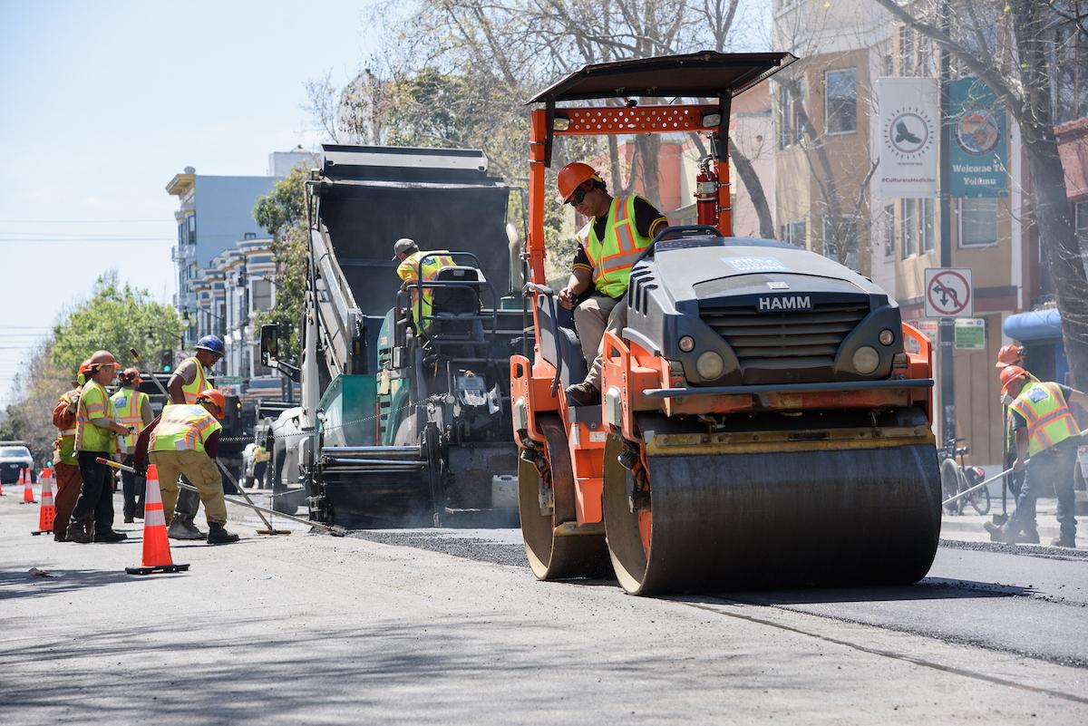 Crews wearing yellow vests repave Valencia Street using large equipment near orange safety cones.