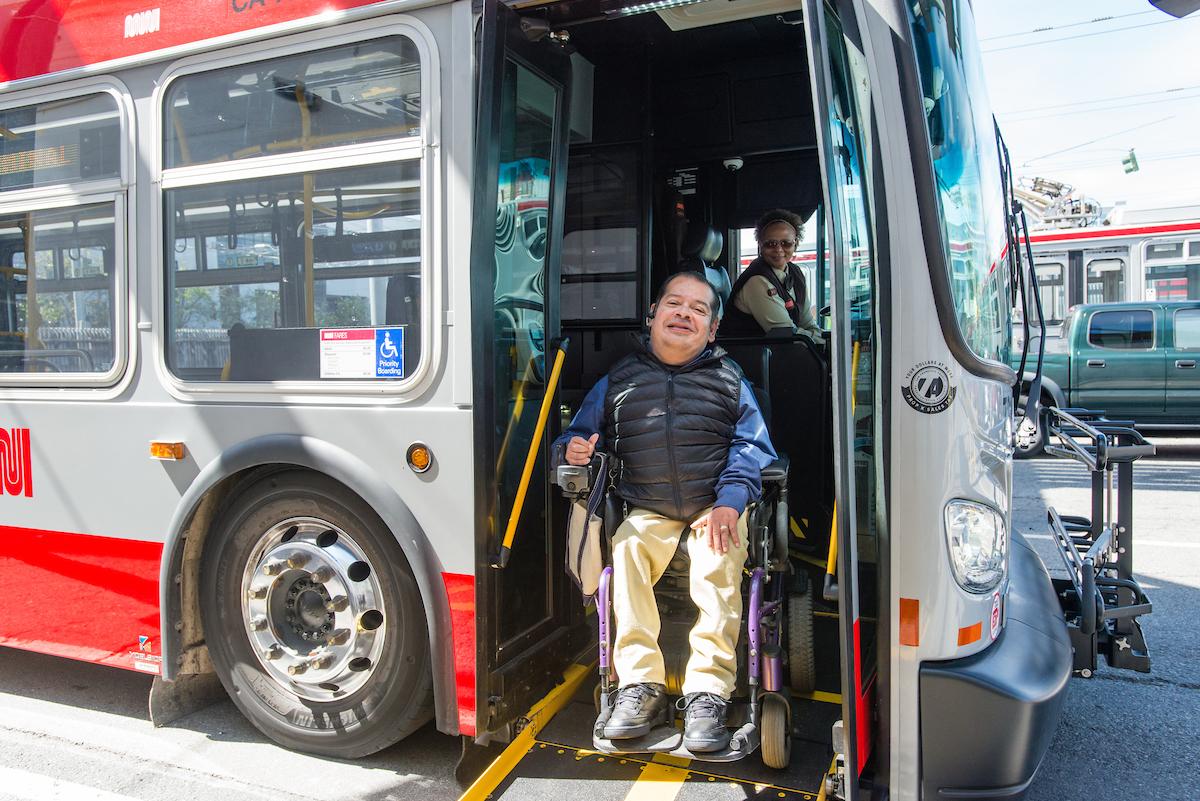 Person using a mobility device exits a Muni vehicle using a ramp as an operator assists.