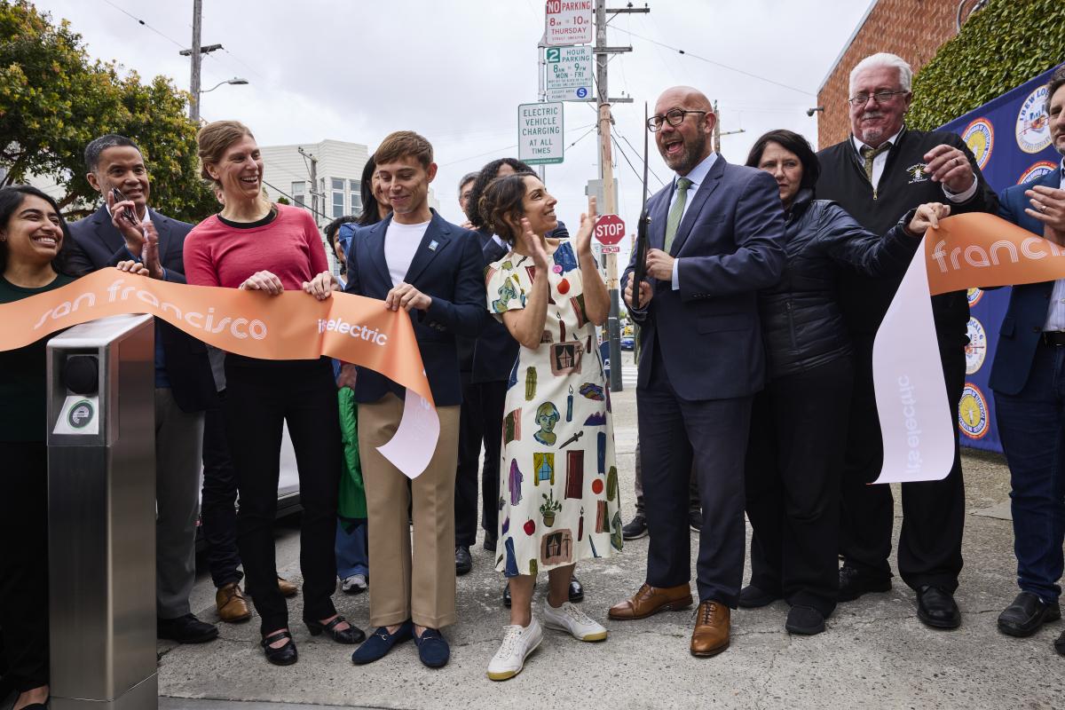Several people smile as they cut a ribbon to celebrate the launch of two curbside EV charging stations.