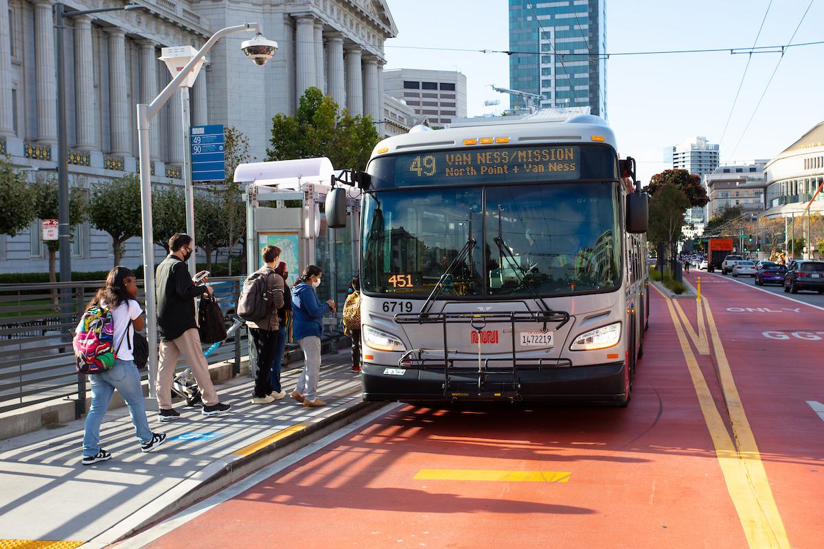 A 49 Van Ness bus stops in a dedicated transit lane in front of City Hall. Several riders line up to board.