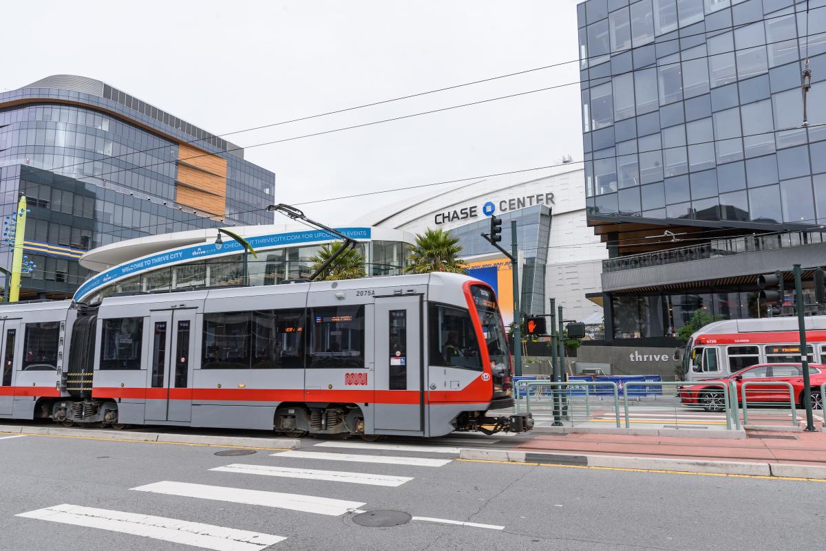 Image shows a T Third Muni Metro train and a 22 Fillmore Muni bus outside of Chase Center in San Francisco.