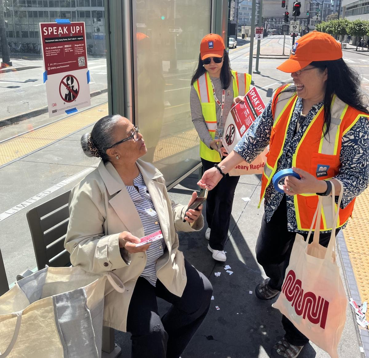 A MuniSafe Day Out volunteer in an orange safety vests speaks to a woman who is sitting down at a Muni bus stop and hands out information to her.