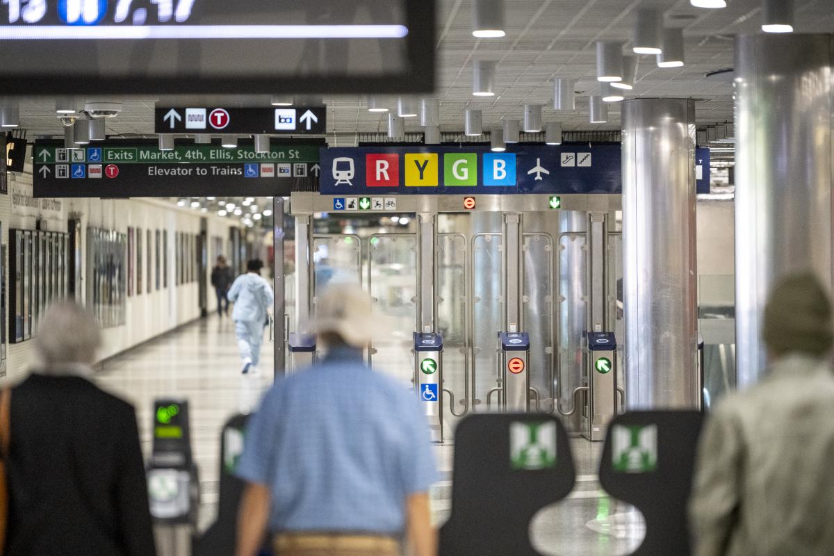 A photograph of transit riders exiting the Muni fare gates at Powell Station with a test sign of the Regional Mapping and Wayfinding Project visible overhead.