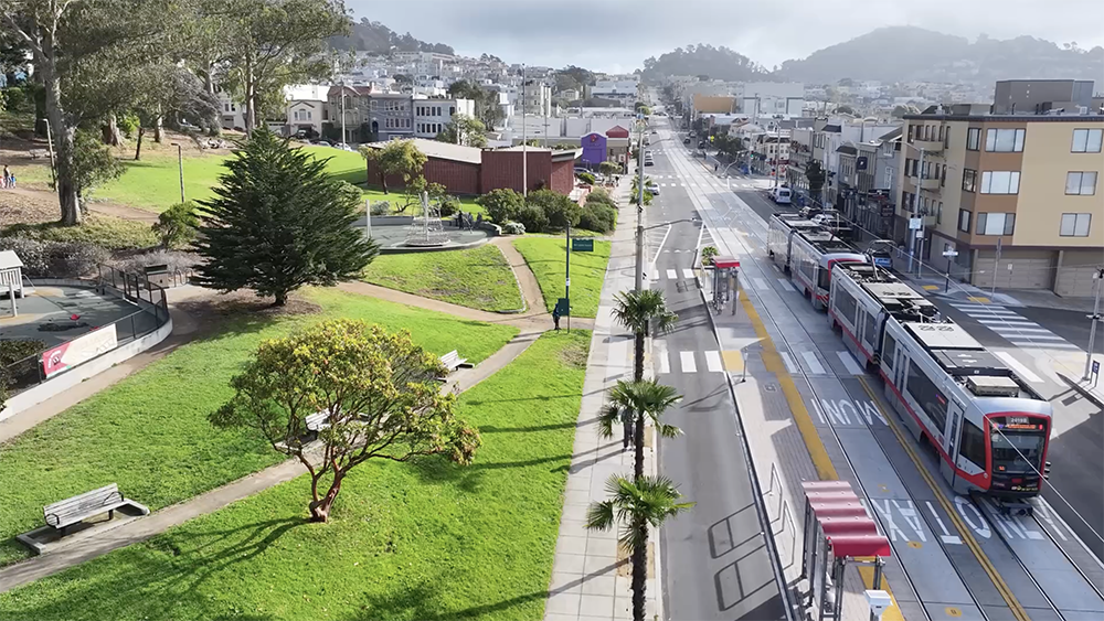 Aerial view of Taraval Street with an L Taraval train passing by a green space with walking paths and trees.