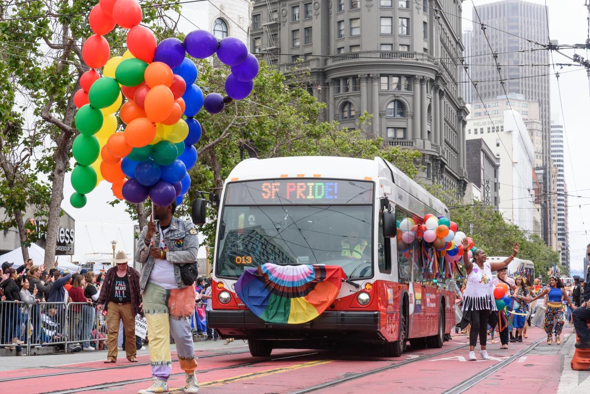 At last year's Pride Parade, a red and white Muni bus that says "SF Pride" and features a rainbow flag drives down Market Street. 