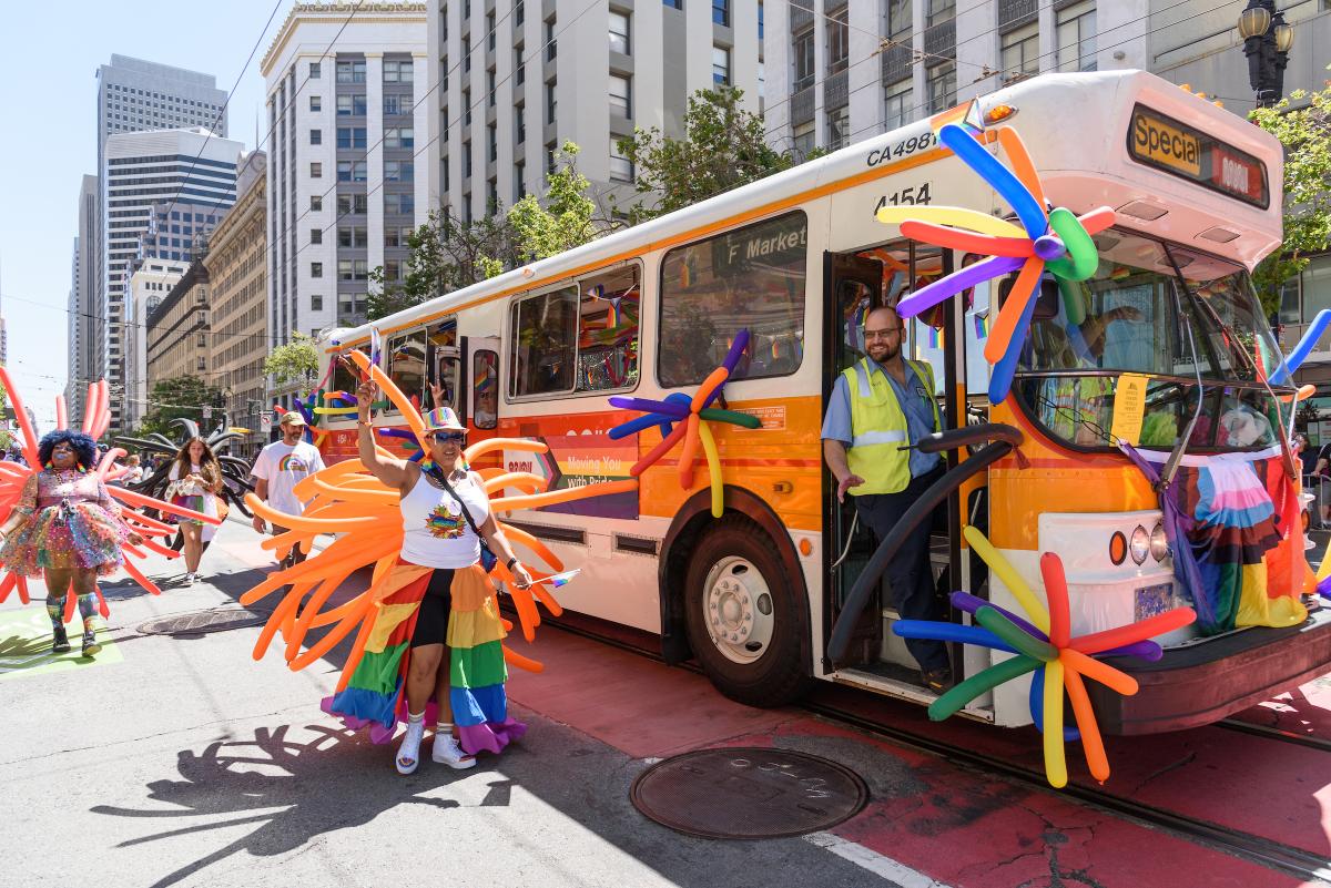 Cream and orange Muni bus drives in the 2024 Pride Parade as people walk by holding rainbow balloons.