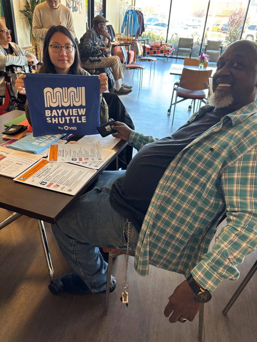 Two people sitting at a table in a commercial space smile as one holds up a tote bag that says Bayview Shuttle.