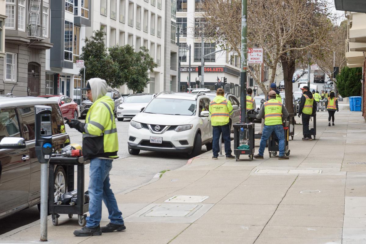 Several crew members in yellow safety vests work to upgrade a few parking meters on a city street.