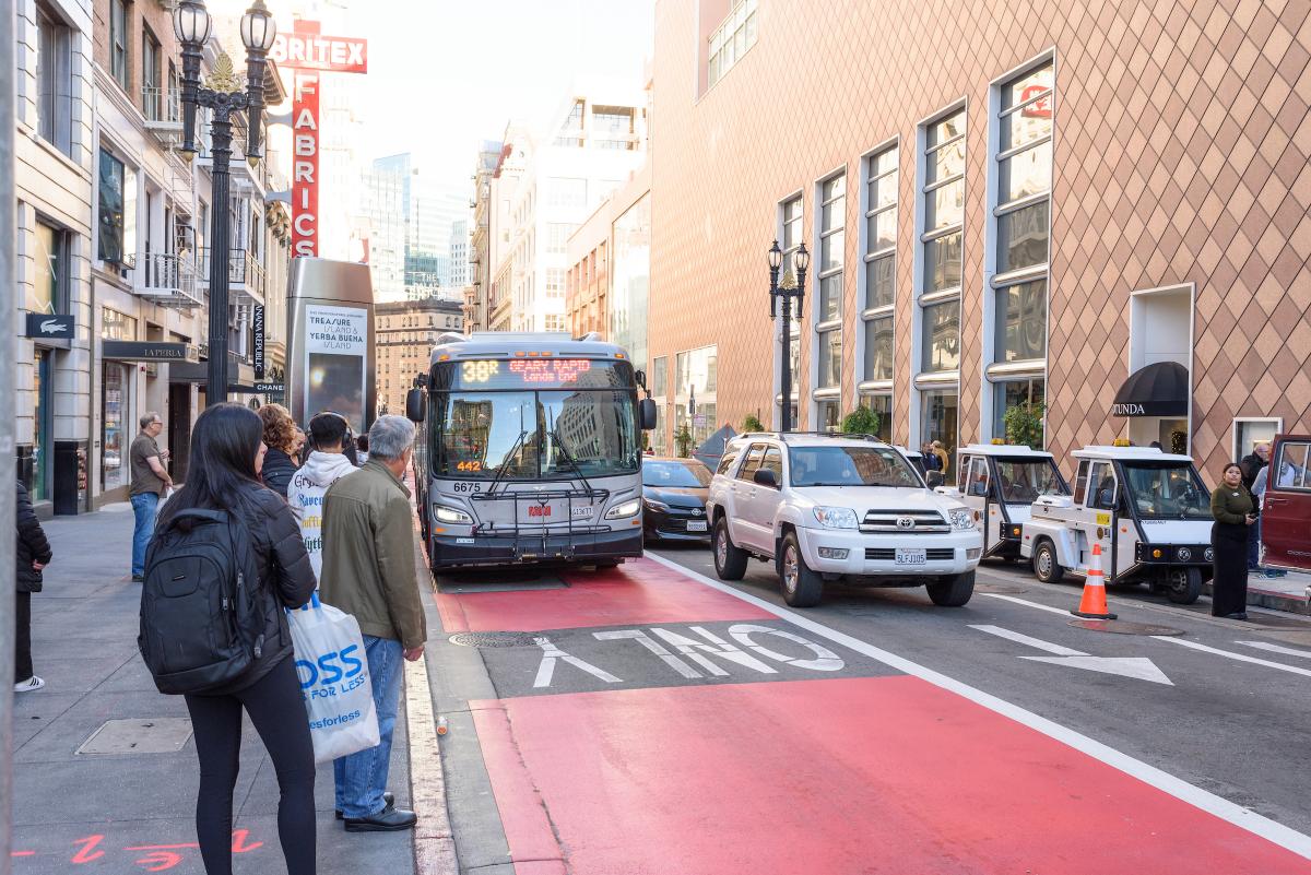 38R Geary Rapid drives in a red dedicated transit lane as people wait on a nearby curb to board.