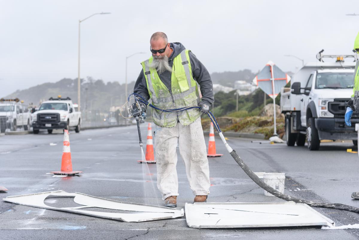 Crew member wearing a yellow vest sprays a lane marking as part of the prep for the Great Highway closure.