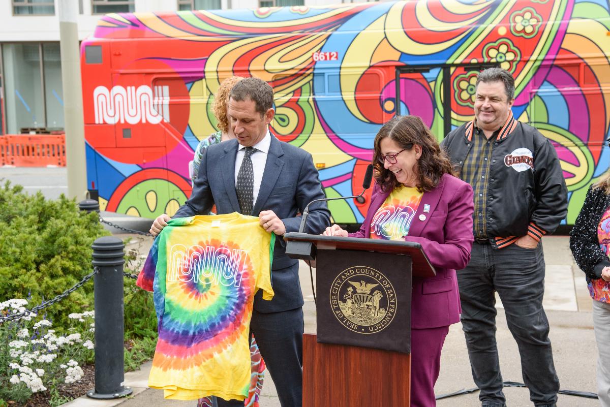SFMTA Director of Transportation Julie Kirschbaum speaks from a podium near Mayor Daniel Lurie, who holds a special Muni-themed tie-dye shirt.