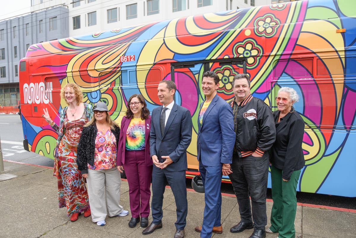 SFMTA Director of Transportation Julie Kirschbaum, Mayor Daniel Lurie and others smile for a photo in front of a bus wrapped in psychedelic colors.