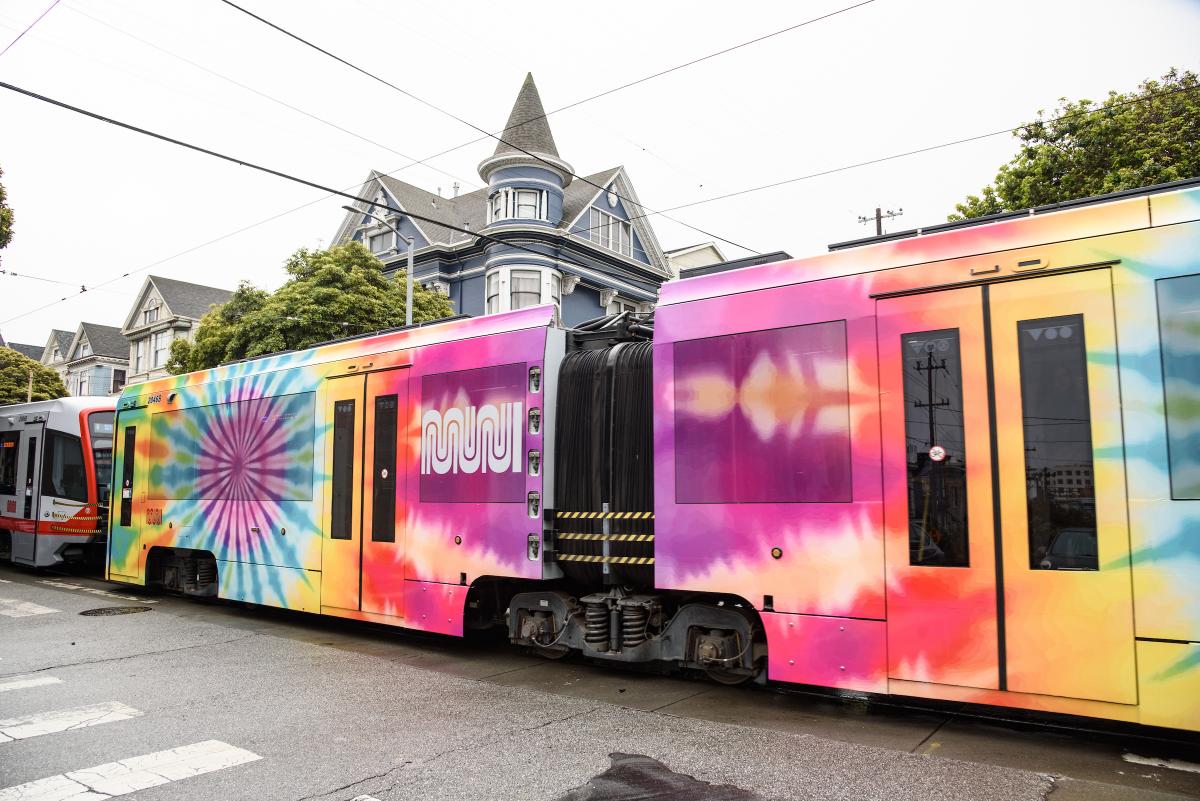 N Judah train wrapped in psychedelic colors for upcoming Golden Gate Park concerts passes an intersection near the Haight.