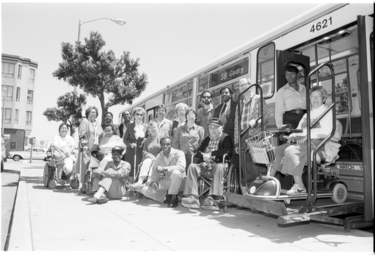 Black and white photo of a group of people posing in front of a Muni bus. Group includes Accessibility Advisory Committee members with Muni staff. 