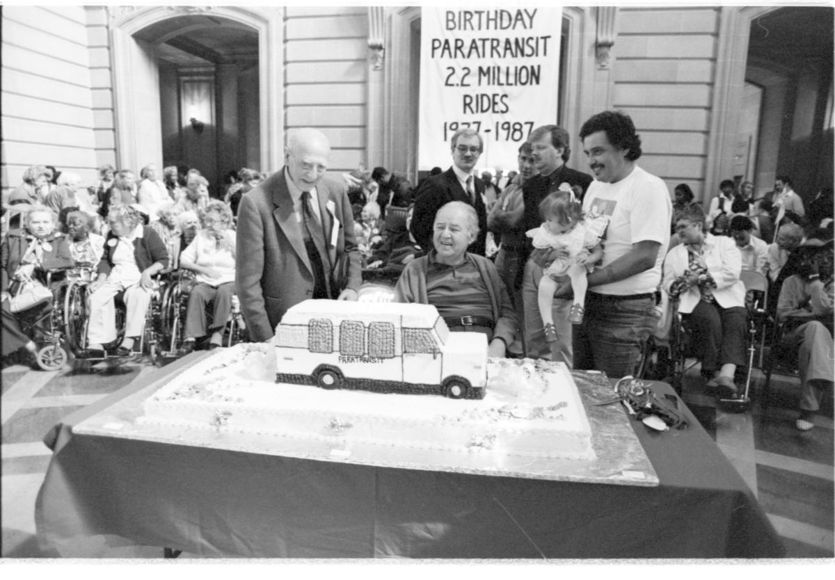 Black and white photo of a group celebrating the 10-year anniversary of paratransit. A small crowd stands in front of a festive cake shaped like a paratransit vehicle.