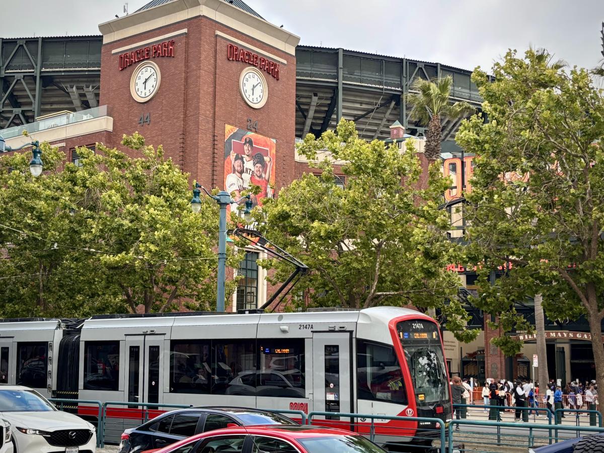 A train in front of a baseball park with people on the sidewalk.
