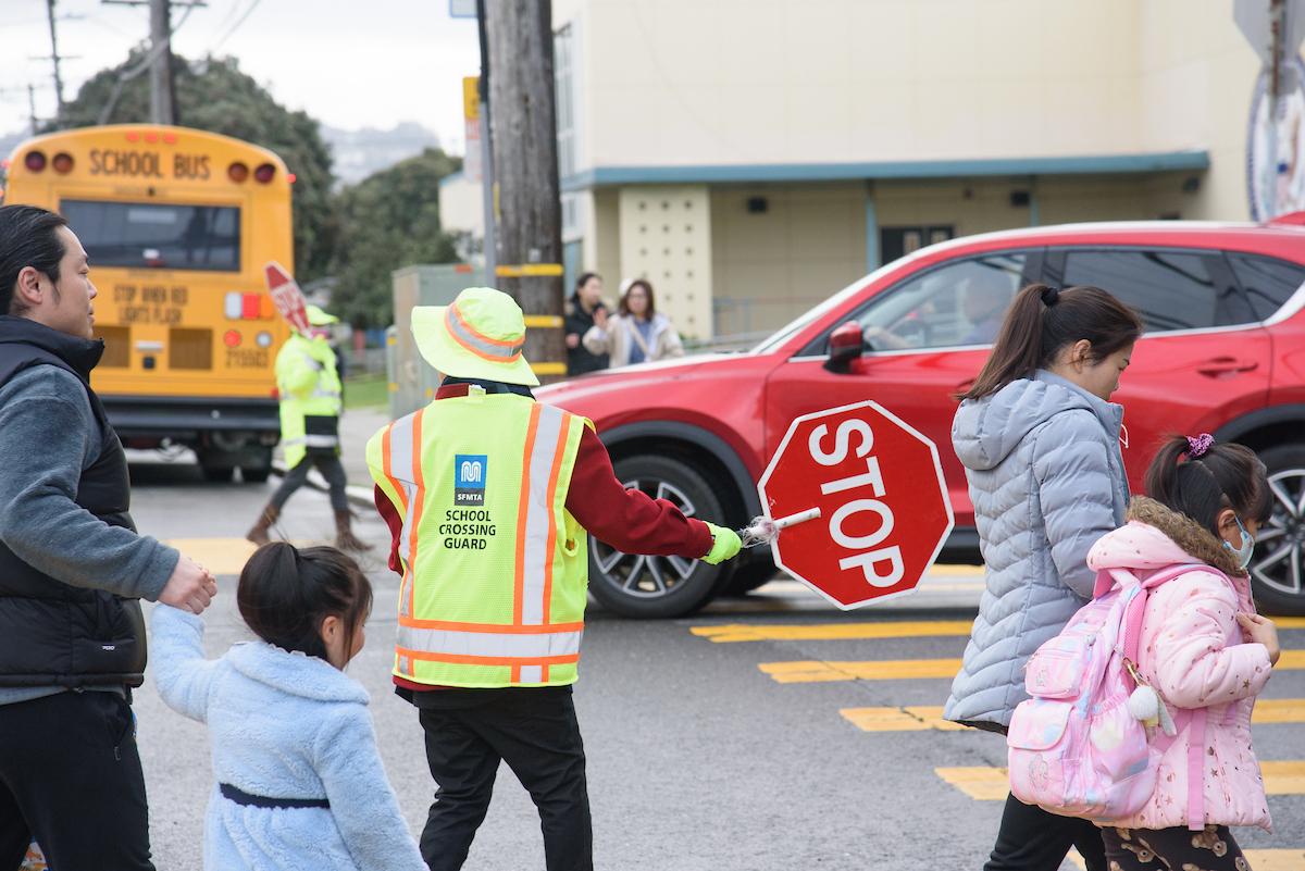 An adult wearing a safety vest and carrying a stop sign in a crosswalk with children and other adults.