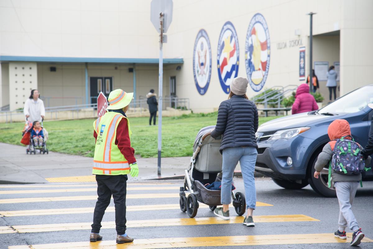Crossing guard in yellow vest helps students and a parent pushing a stroller cross the street.