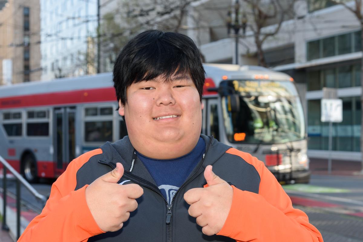 Peter, an Asian young man black hair, is pictured smiling on the street in front of a Muni bus with a “thumbs up” on both hands. He wears an orange and black sweater.