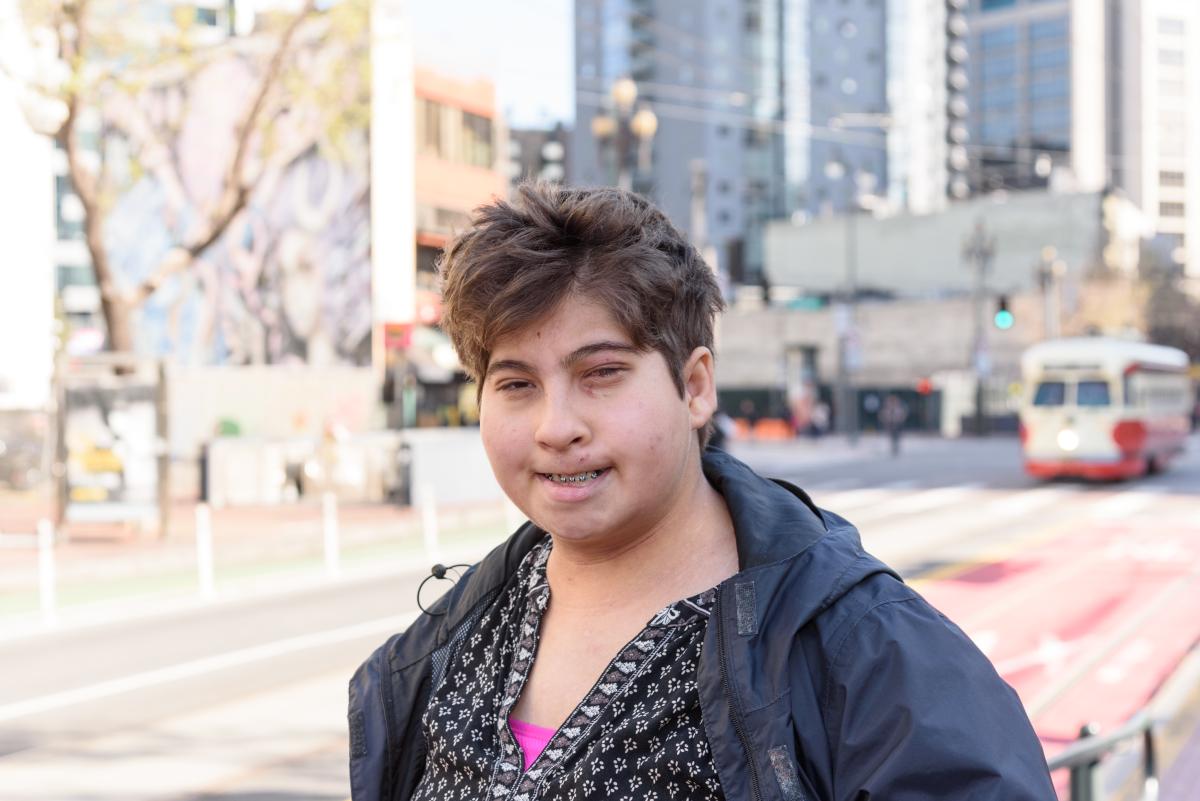 Belen, a young woman with light skin and short brown hair, is pictured smiling on the street in front of a Muni trolley.  She wears a black shirt and navy hoodie.