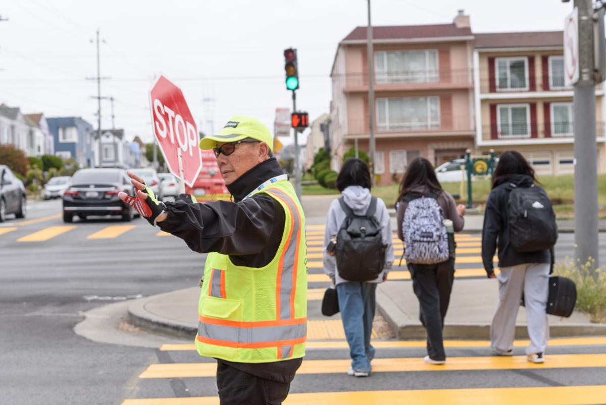 Crossing guard wearing yellow vests helps three students cross the street.