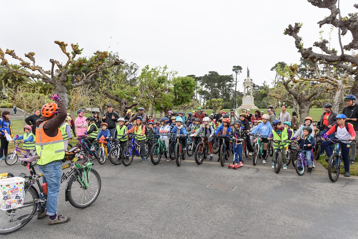 Children and some adults waiting on bikes in a park.