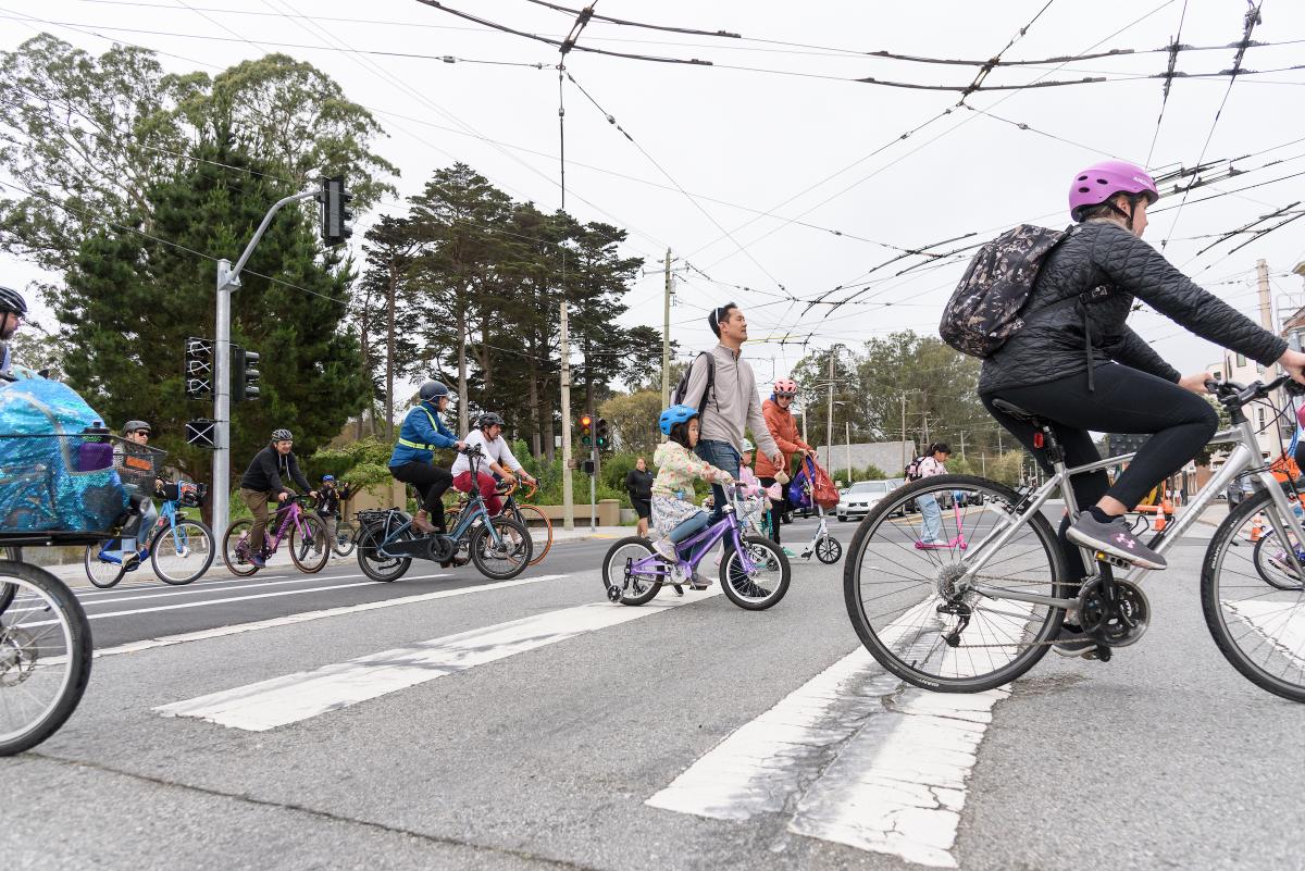 Adults and young children bike across a street near a park.