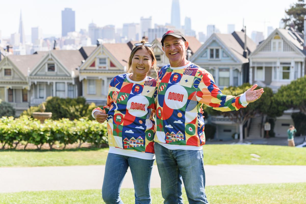 Two people wear the Patchwork of the City Holiday Pullover at Alamo Square in front of the Painted Ladies.