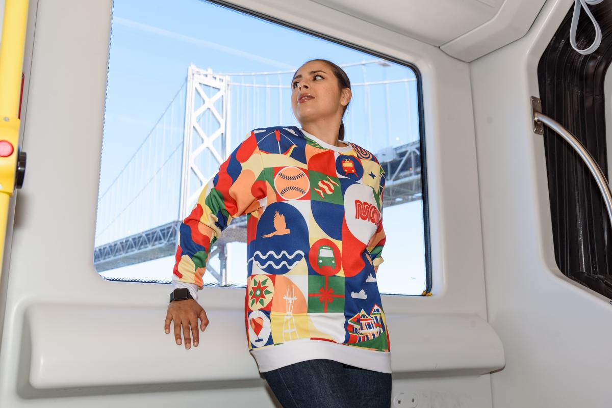 Person wears the Patchwork of the City Holiday Pullover on a Muni Metro train with the Bay Bridge seen in the background of the photo.