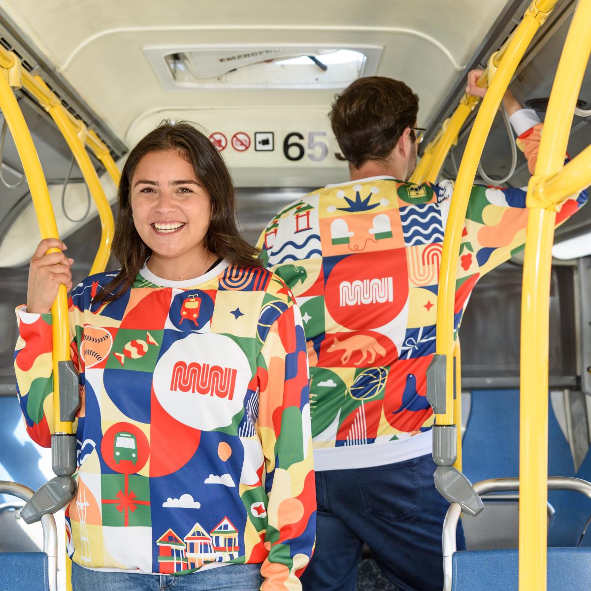 Two people wearing our new Patchwork of the City Holiday Pullover smile on a Muni bus. One faces the camera and the other is turned away, showing the back of the sweater.