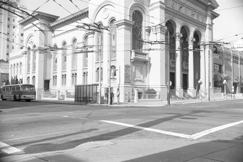 A black and white photo of a large white building and a vintage bus parked outside.