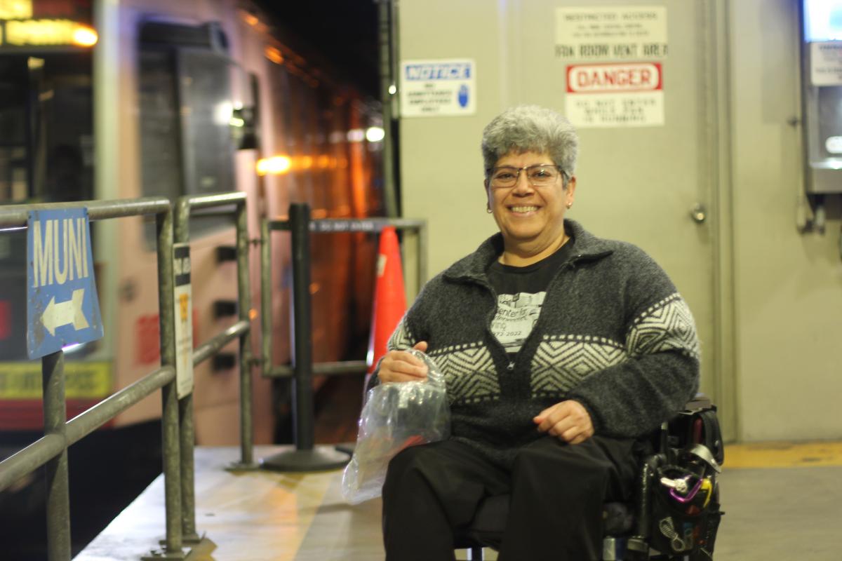 Alicia, a gray-haired Latine woman, is pictured in a power wheelchair on a Muni metro train platform as the train enters the background.  She wears a grey sweater and glasses.