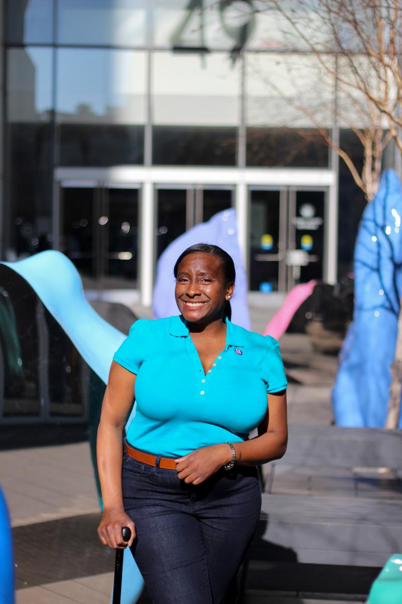 Ammy J Ammy, a Black woman is pictured smiling in a sculpture garden wearing a bright blue top and jeans. She holds a cane in her righnt hand.