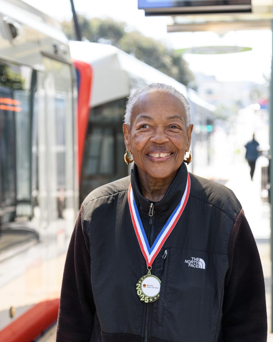 Claire, a gray-haired Black woman is pictured smiling with a medal around her neck. She is wearing a black jacket.  A metro vehicle passes behind her.