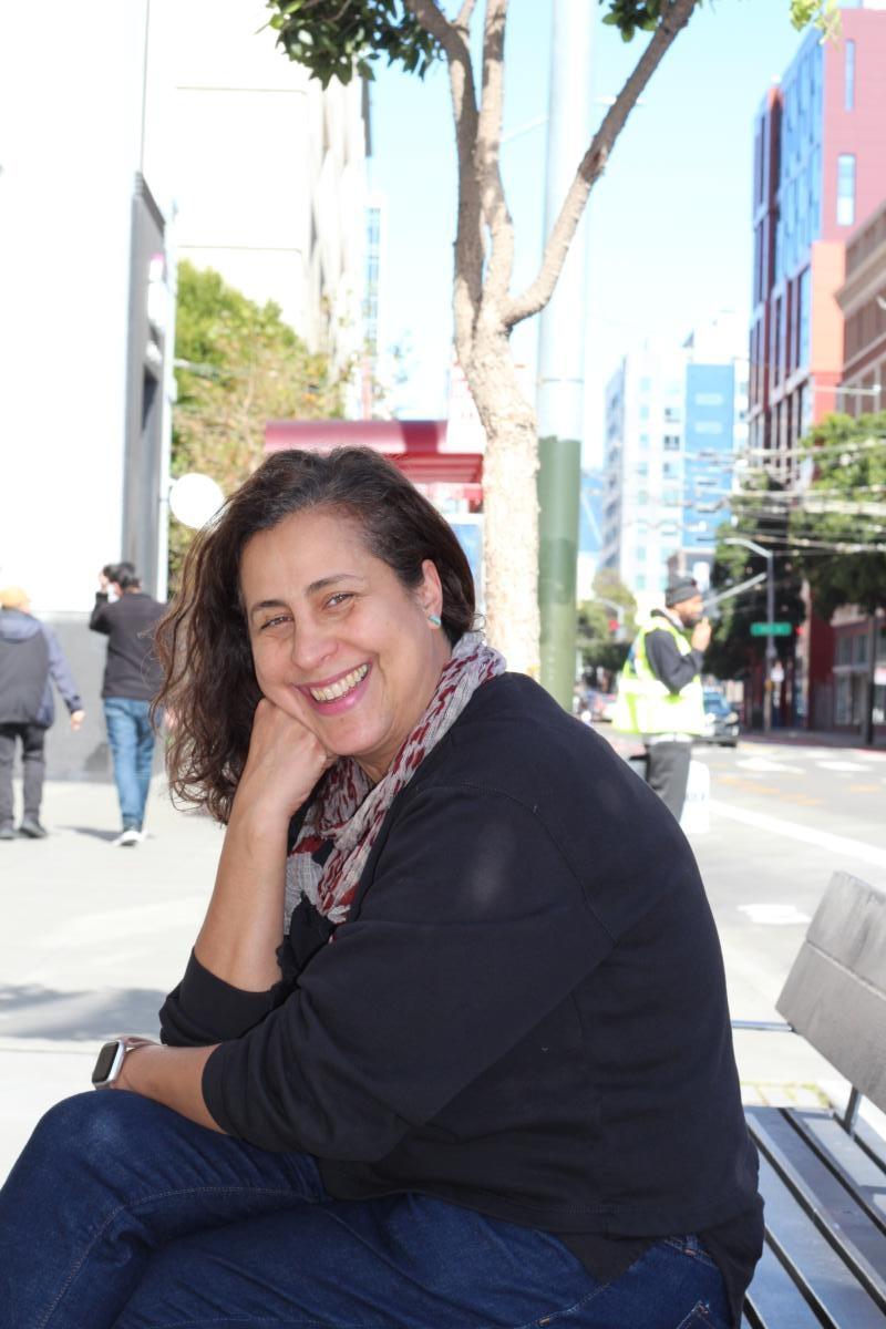 Orkid  is pictured sitting on a bench, smiling while seated on a bench in front of a Muni bus shelter. 