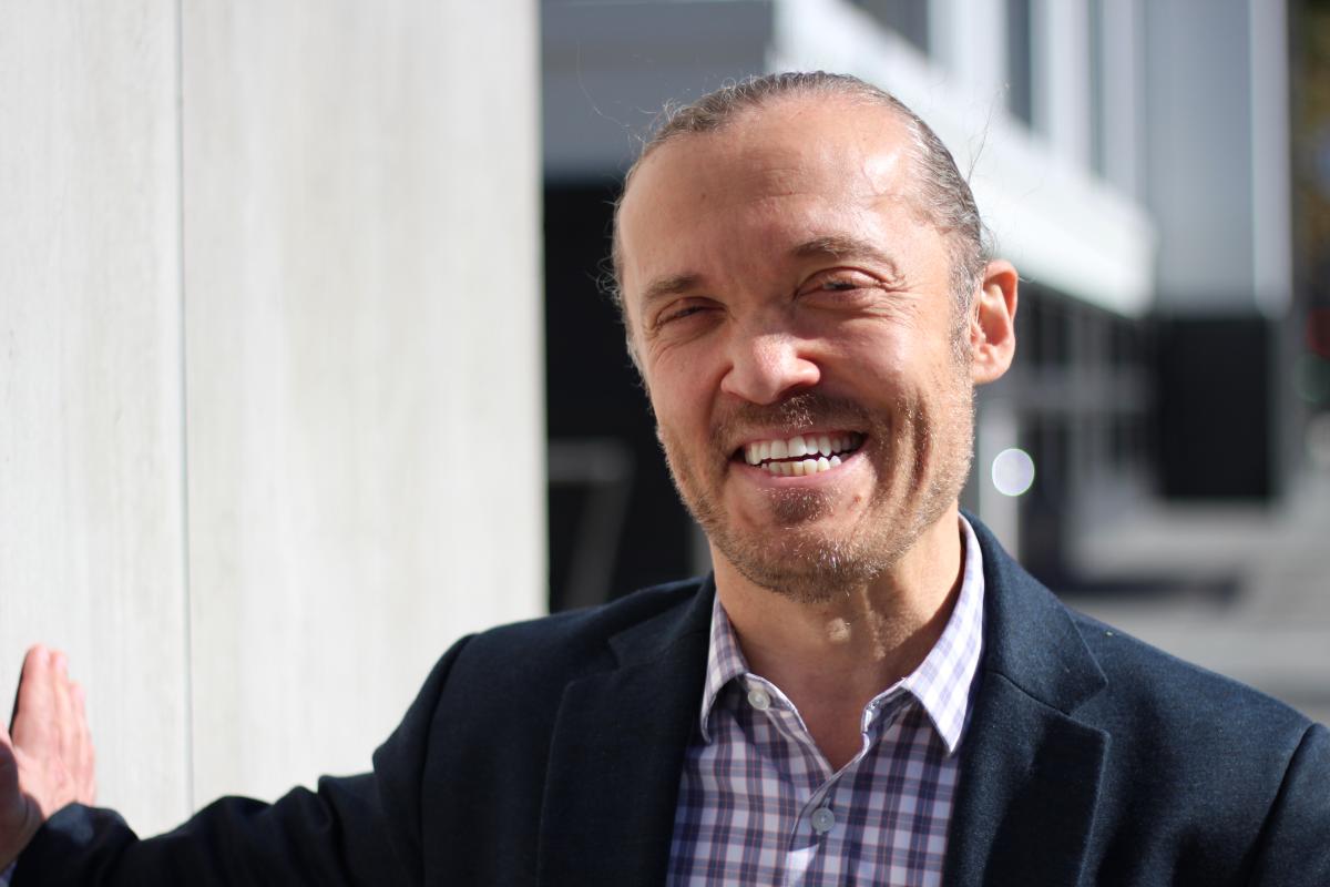 Eli, a White man slicked-back hair, is pictured outdoors smiling and wearing a suit