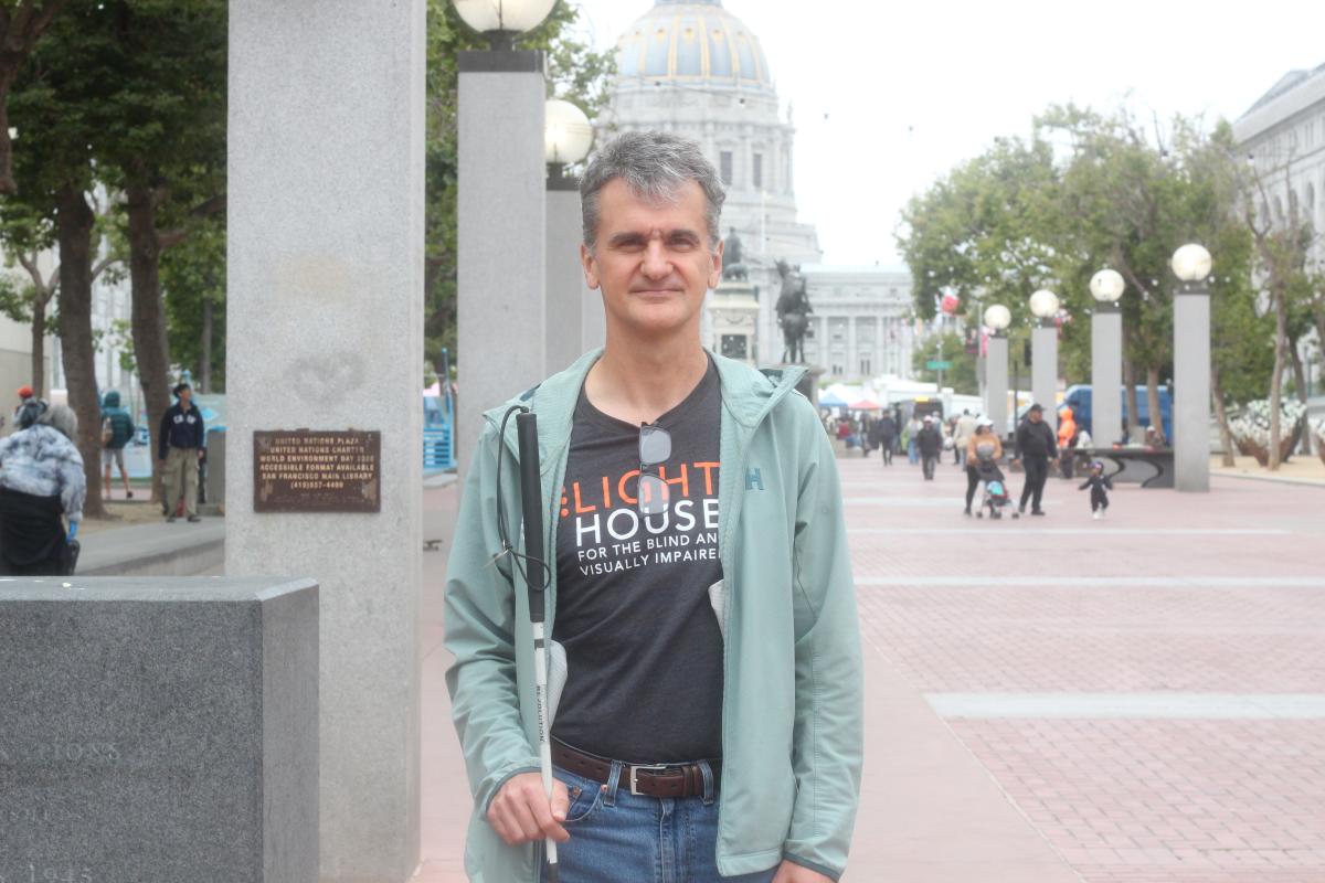 Corvin faces the camera directly.  He wears a dark grey Lighthouse for the Blind t-shirt with his glasses tucked in the collar and a light green jacket.City hall can be seen in the background. He holds a blind cane.