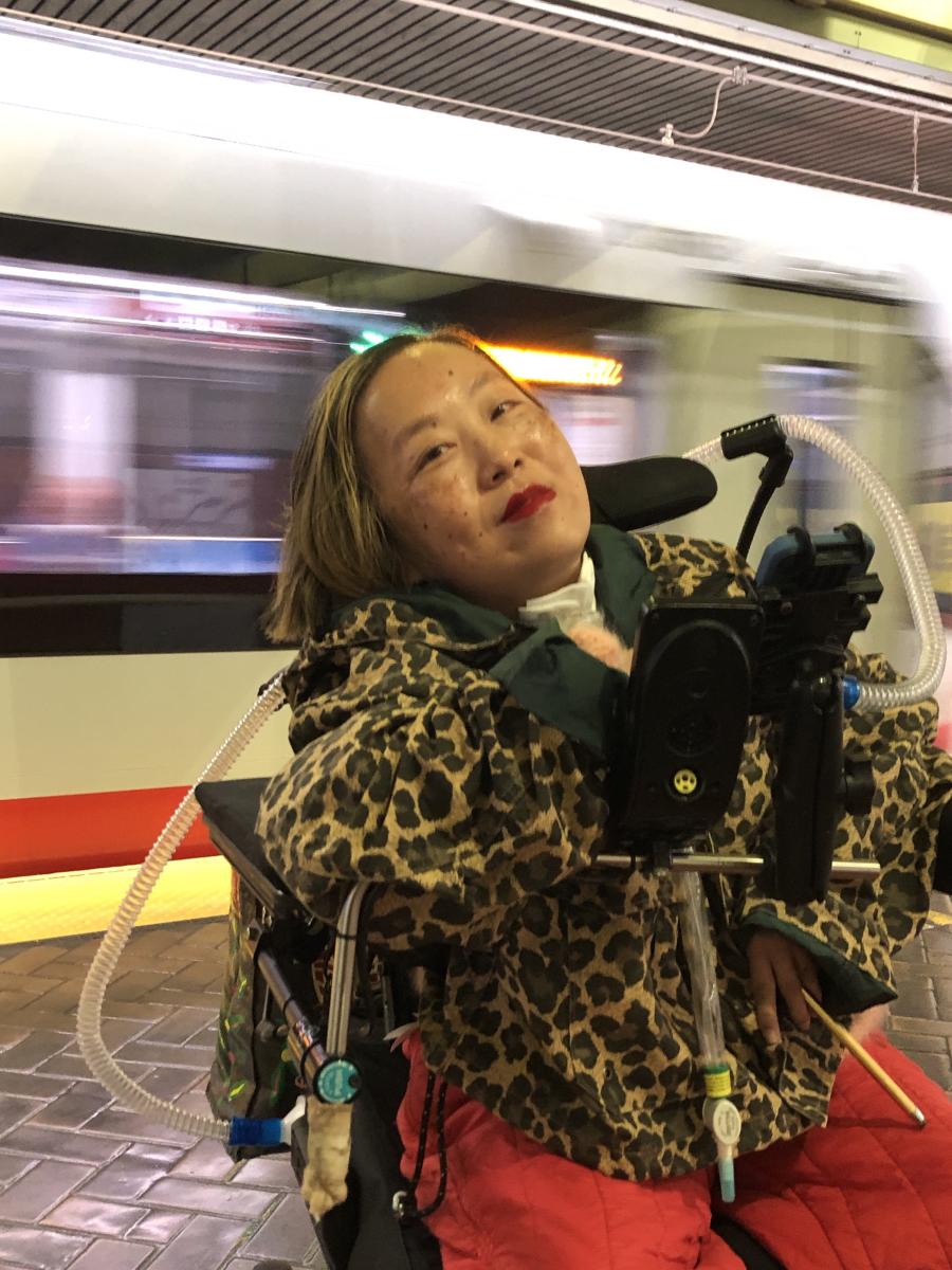Alice, an Asian American woman with a tracheostomy at her neck is pictured smiling inf front of a moving Muni train. She is wearing red lipstick and leopard print jacket.