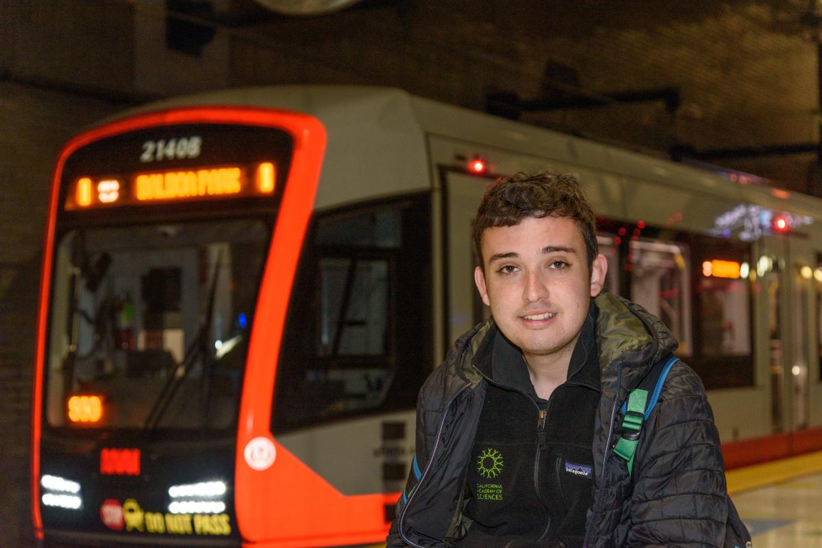Julian Kanter Julian Kanter, a young White man, is pictured with a neutral expression in front of a Muni train as it arrives. He is wearing two black jackets and a backpack