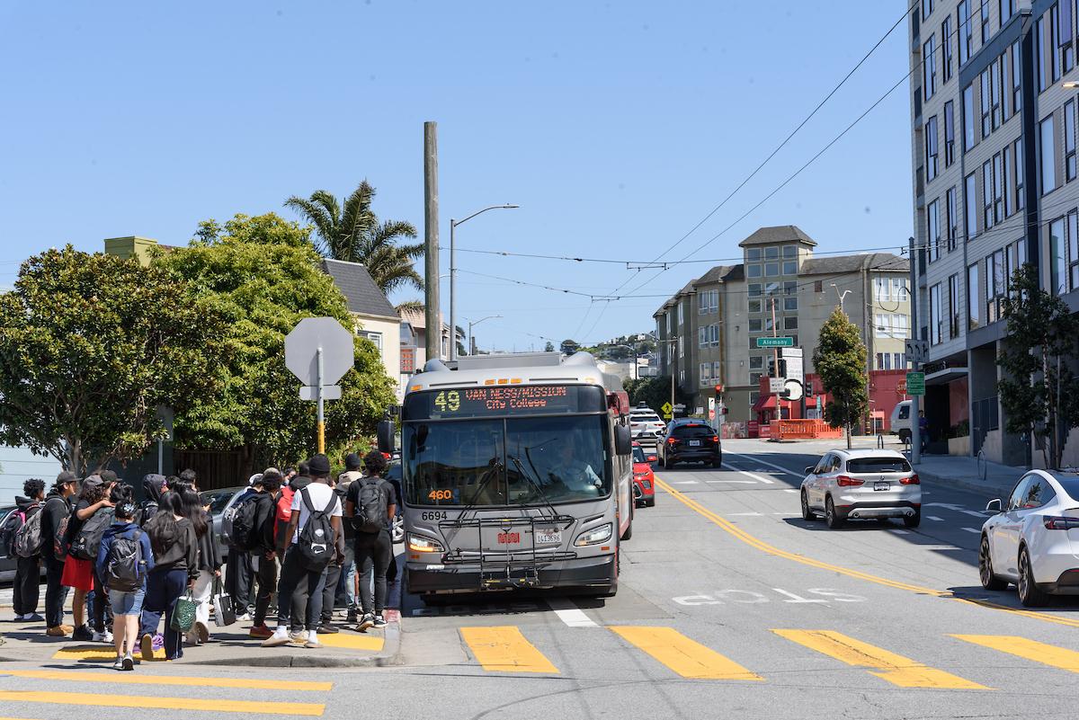 Several young people wait near a corner to board a 49 Van Ness.