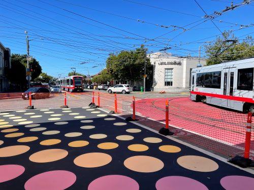 Outside of West Portal Station, trains pass a newly painted street mural of a black background with multicolored dots.