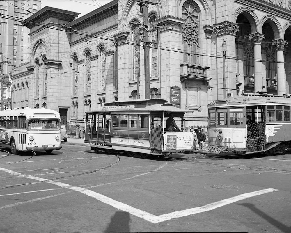 A black and white vintage photo of a cable cars, a vintage streetcar, and a retro bus at an intersection in front of a stone building.