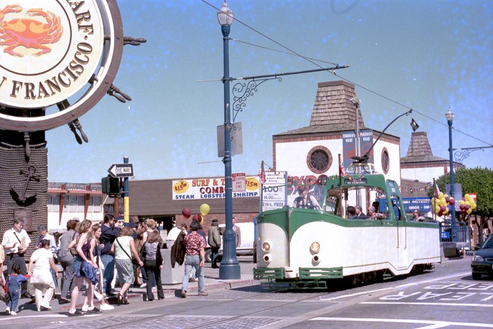 Green and white "Boam Tram" historic streetcar drives near the piers as people walk on a nearby sidewalk.