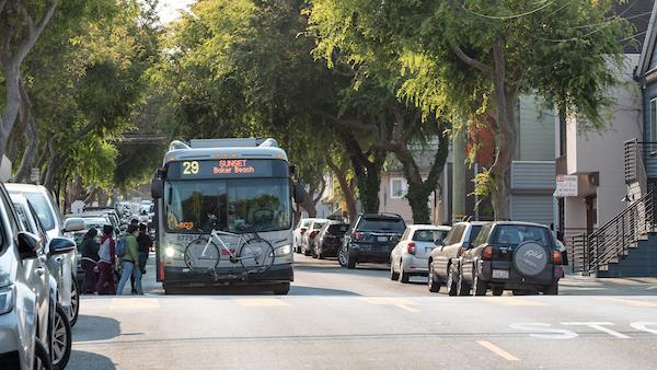 A 29 Sunset Bus stops on a tree-lined street where several people board near parked cars.
