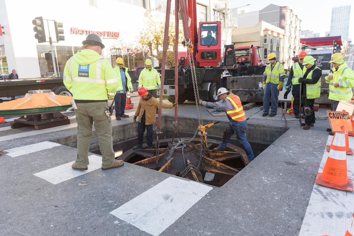 Large group of people wearing safety vests standing near a crane as it lefts a cable car sheave out of the ground for restoration work.