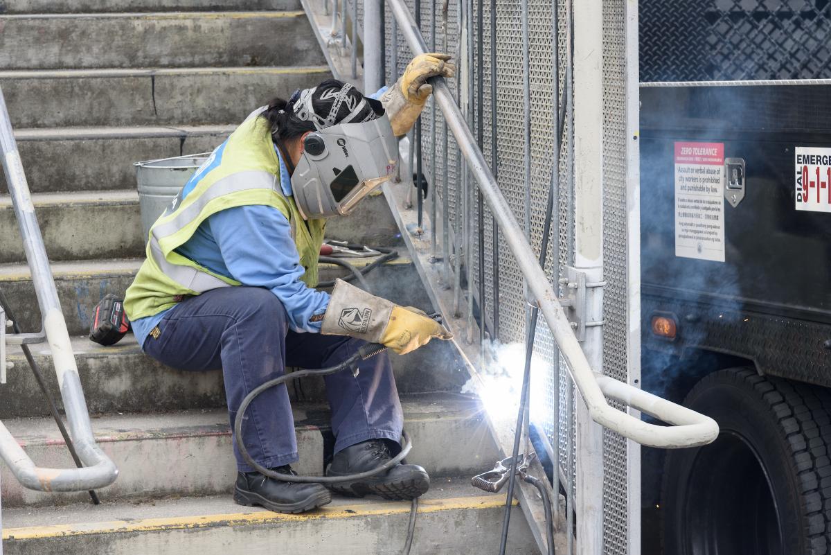 Person wearing yellow safety vest uses a welding tool to repair a staircase railing for our first Fix-It Weekend.