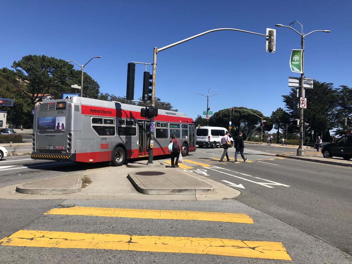 44 bus drives through an intersection as people walk in a crosswalk nearby.