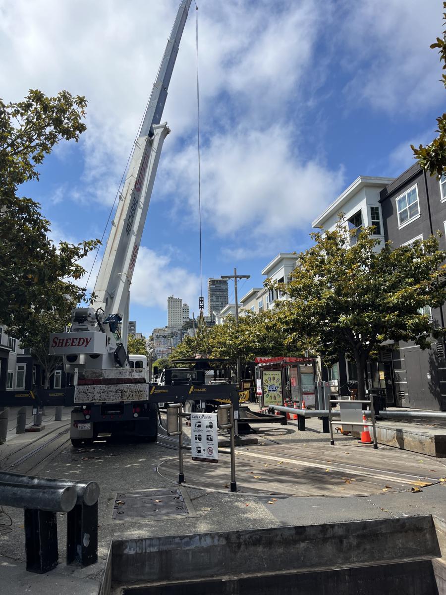 A large crane lifts a refurbished sheave from the ground against a bright blue sky.